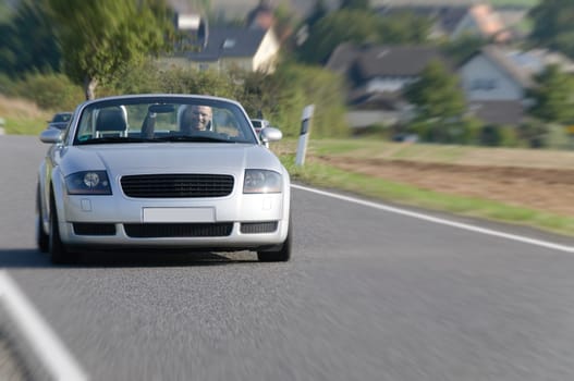 Young man in a suit driving fast in his open silver sports car on a country road.