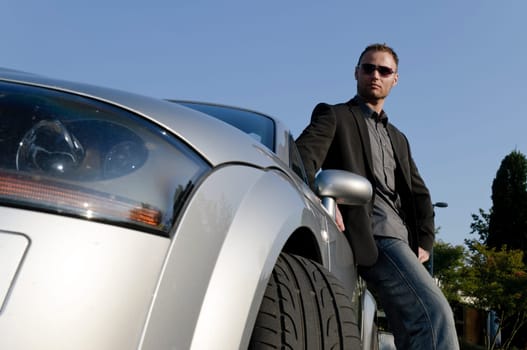Young man in a suit stands confidently in front of his silver open-topped car.