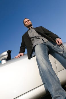 Young man in a suit stands confidently in front of his silver open-topped car.