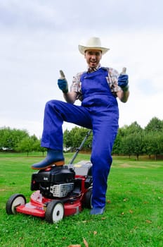 Adult man in blue overalls (gardener), rubber boots and straw hat sitting proudly with his arms crossed on his lawnmower in the background you can see a large grassy plot
