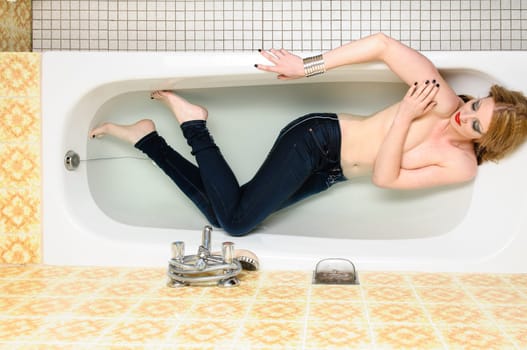 Beauty blond young woman lying with her bluejeans in the bathtub in water.