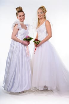 Two young women wearing bride dresses und holding their bride bouquets, against white background. Smiling in to camera
