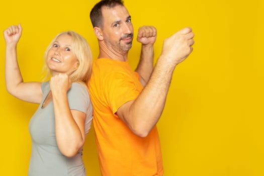 Back to back happy caucasian couple in their 40s celebrating victory with hands in the air on yellow studio background