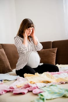 Pregnant woman with headache above baby clothes at home on sofa