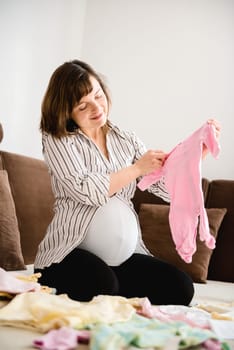 Pregnant woman preparing baby clothes at home on sofa