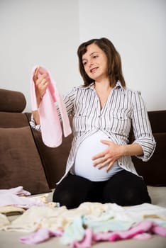 Pregnant woman preparing baby clothes at home on sofa