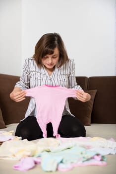 Pregnant woman preparing baby clothes at home on sofa