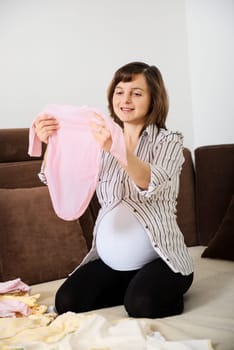 Pregnant woman preparing baby clothes at home on sofa