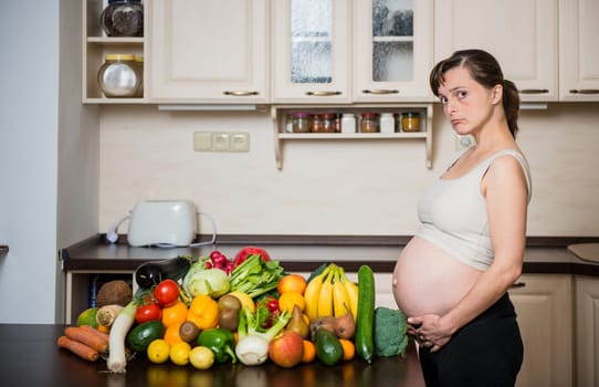 Unhappy pregnant woman with abundance of vegetables and fruits in kitchen