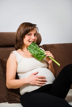 Lifestyle portrait of smiling pregnant woman biting spinach at home