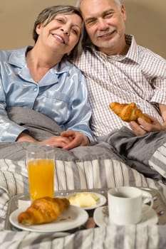 Happy retired couple eating croissant for breakfast in bed