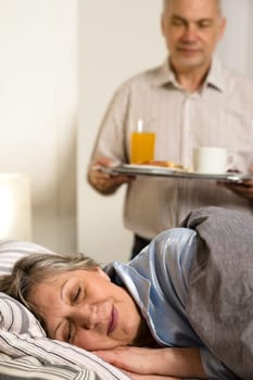 Loving senior husband serving breakfast to sleeping wife