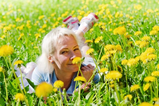 A woman lies in a clearing among the dandelions and sniffs a flower