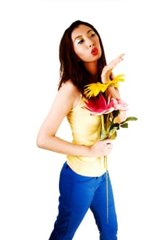 A tall young Chinese woman standing for white background in blue pants and yellow top, blowing a kiss and holding some flowers in her hand.