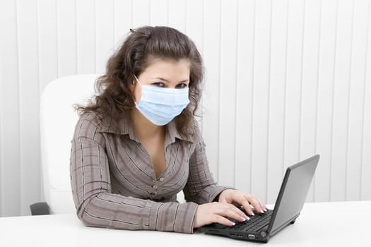 The young woman at office in medical mask works with the laptop