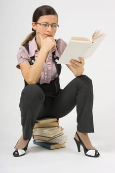 a young woman sitting on a pile of books with glasses and book in hand