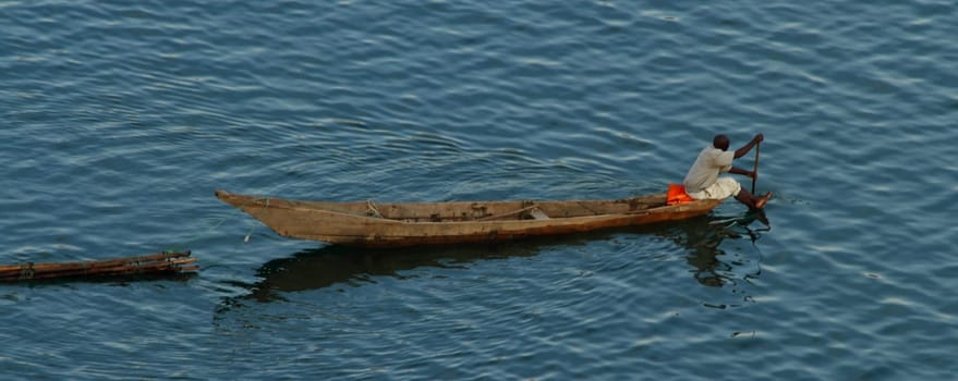 A lone African man paddles his long boat across a lake while pulling some logs.
