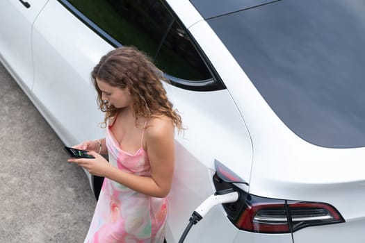 Aerial top view eco-friendly woman recharge electric vehicle from EV charging station, using EV technology utilization for tracking energy usage to optimize battery charging on smartphone. Synchronos