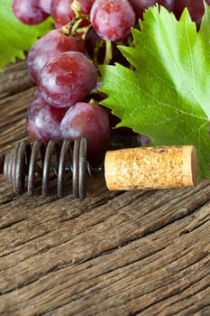 Old corkscrew with a cork lies between red grapes and vine leaves on rustic wooden ground