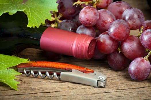 Ripe red grapes with vine leaves lie on a wooden surface together with a wine bottle and corkscrew