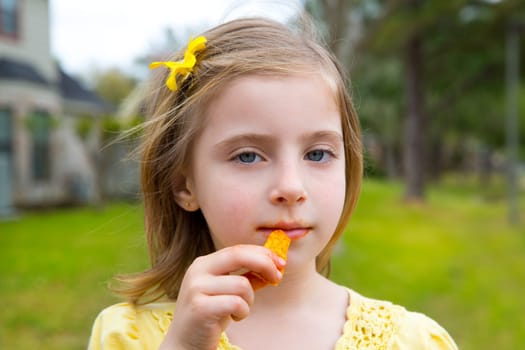 Blond kid girl eating corn snacks in outdoor park in lawn background