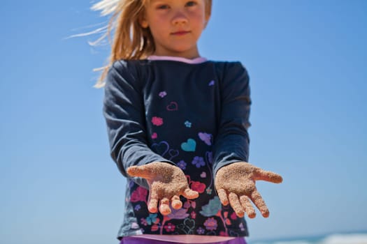 Cute young girl holding out her sand covered hands against blue sky background while playing at beach