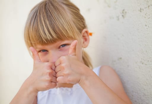 Closeup of cute young girl doing thumbs up ok sign while leaning against concrete wall