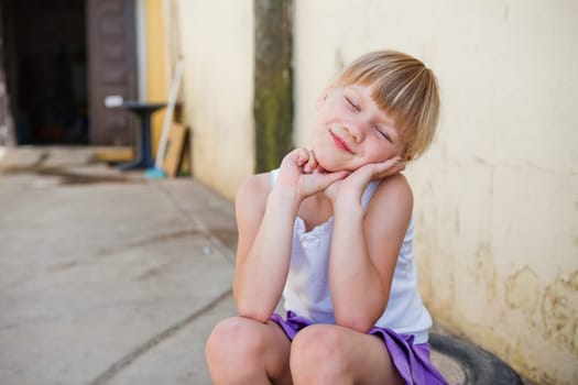 Portrait of cute smiling young girl with eyes closed sitting in front of concrete wall outside