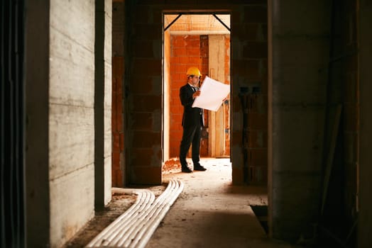 Engineer at work in construction site, standing in apartment building and looking at blueprints and plans