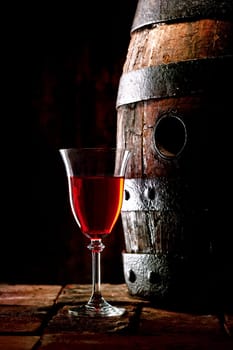 A glass of red wine next to an old oak cask with its stopper out.