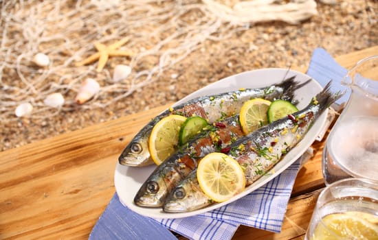 Three grilled sardines on a platter over a table outdoors on the beach with a fishing net and a starfish in the background