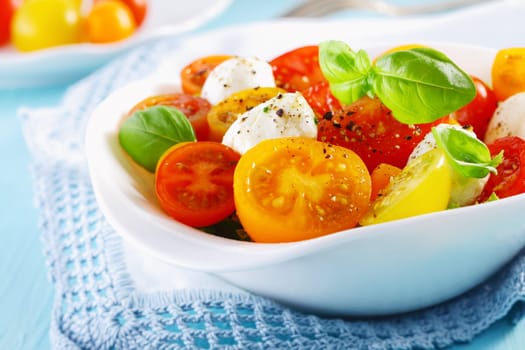 White bowl of fresh and healthy Mediterranean salad with mozzarella cheese, tomatoes and basil leaves over a white doily blue table