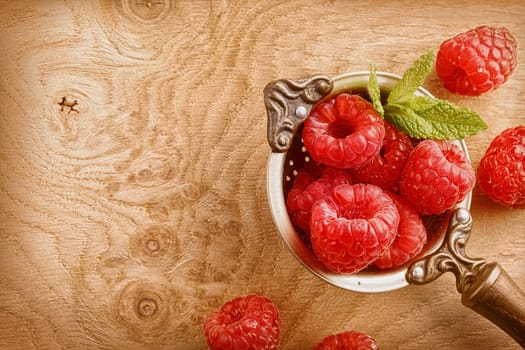 Top view of an old copper ladle filled with ripe raspberries decorated with mint leafs over a wooden table