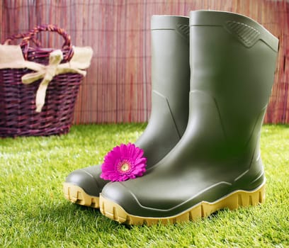 Gumboots with a pink gerbera daisy standing on a neat green lawn in front of a rustic wicker basket in a gardening and healthy lifestyle concept