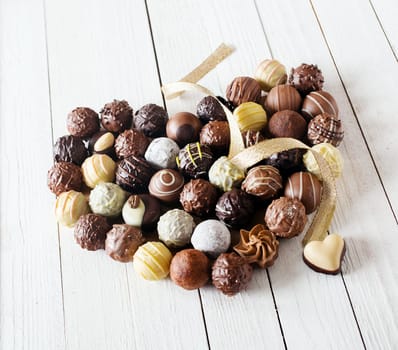 Close-up of a heart shape made with various types of chocolate truffles decorated with a curly golden ribbon over a white wooden table