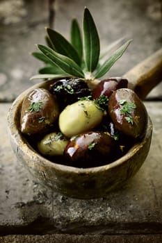 Close-up of an old wooden ladle or large spoon filled with seasoned black and green olives decorated with an olive-tree branch resting over a stone