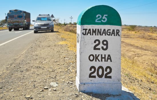 Horizontal color landscape of the 29 kilometers to Jamnagar direction milestone of the Gujarat state highway in India. Stationary vehicle and truck whizzing past under clear blue sky up to the horizon line