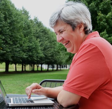 middle-aged woman browsing on laptop at table outside with trees