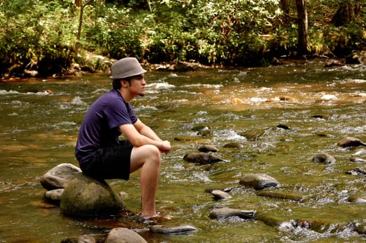 Teenager sitting on river rocks, with hat on, side view