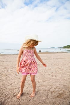 Thoughtful young barefoot girl wearing hat and pink patterned dress at beach with ocean in background