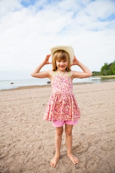 Smiling young barefoot girl wearing hat and pink patterned dress at beach