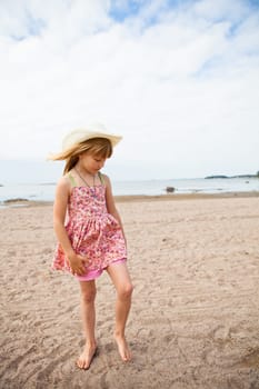 Thoughtful young barefoot girl wearing hat and pink patterned dress at beach with ocean in background