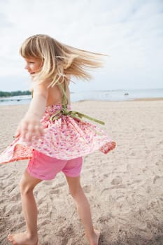 Smiling young barefoot girl with pink patterned dress and pants having fun at beach with ocean in background