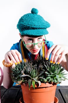 Young woman with black straight hair wears colorful mixed clothes and stands in front of a white background and devotedly pours a cactus.