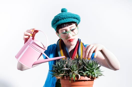Young woman with black straight hair wears colorful mixed clothes and stands in front of a white background and devotedly pours a cactus.