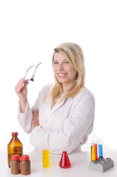 Attractive young chemists with blond hair and glasses wears a white coat. She stands contentedly and proudly with folded arms behind a table with various chemicals,exposed against a white background