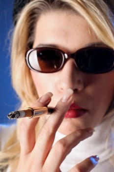 Attractive young woman with long blonde hair wears a tight sleeveless turtleneck top,black shorts with suspenders and high heels. She casually smokes a cigarette with a cigarette tip,against a dark background.
