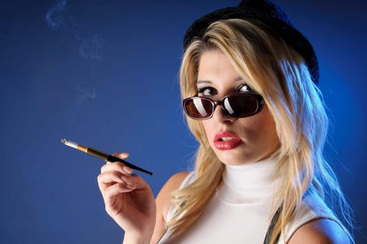 Attractive young woman with long blonde hair wears a tight sleeveless turtleneck top,black shorts with suspenders and high heels. She casually smokes a cigarette with a cigarette tip,against a dark background.