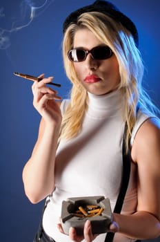 Attractive young woman with long blonde hair wears a tight sleeveless turtleneck top,black shorts with suspenders and high heels. She casually smokes a cigarette with a cigarette tip,against a dark background.