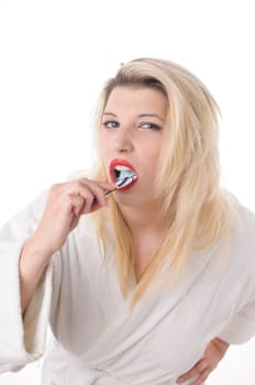 Attractive young woman with long blonde hair wears a white bathrobe and brushes her teeth with a toothbrush,exposed against a white background.